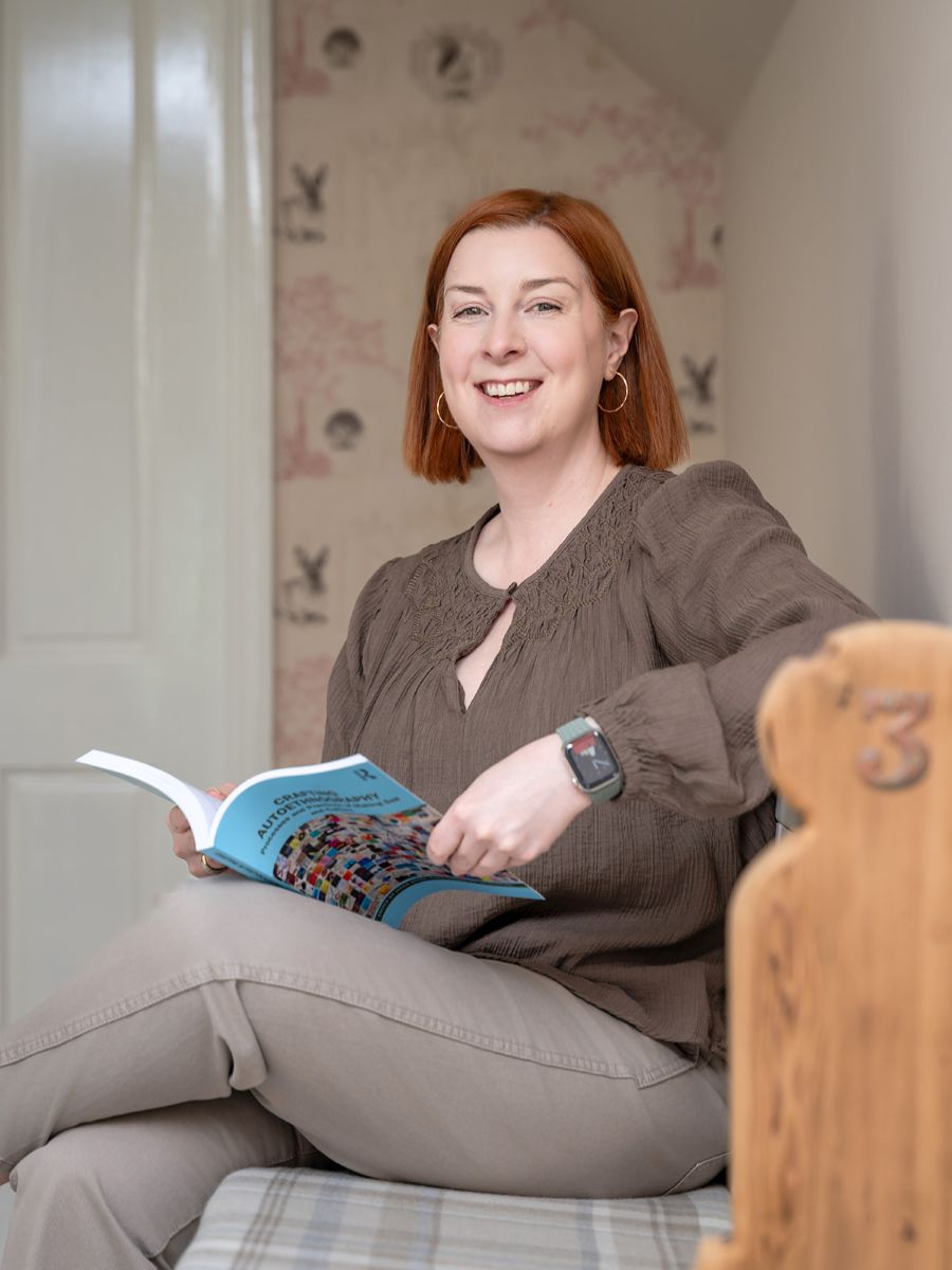 About Dr Karen Lumsden - white woman with strawberry blonde hair, wearing greeny-blue top, sitting at a kitchen table with a blue mug in her hands.