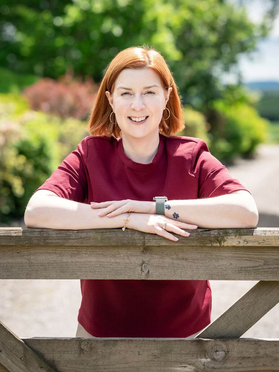 About Dr Karen Lumsden - a black and white photo of a white woman wearing jeans, a blue top and jacket, sitting on a chair with one leg underneath her, holding an ipad.